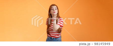 Silly cute and flirty redhead passionate girl, send air kiss camera, fold lips hold arm near mouth to blow mwah, romantic tender attitude, confess love like guy, stand orange background Silly cute and flirty redhead passionate girl, send air kiss camera, fold lips hold arm near mouth to blow mwah, romantic tender attitude, confess love like guy, stand orange background 98145949