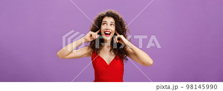Woman waiting entertained for loud bang and fireworks smiling broadly gazing at upper right corner covering ears with index fingers standing charmed in luxurious red dress and evening makeup 98145990