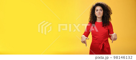 Portrait of gloomy girl with sad face pursing lips down frowning disappointed and pointing down with regret and unsatisfied expression, missing opportunity feeling upset over yellow background 98146132