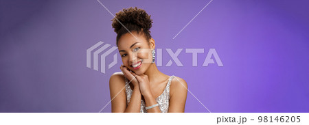Tender elegant wealthy young african american rich woman in brilliants silver stylish shiny dress lean head shoulder smiling flirty romantic gaze camera standing amused blue background 98146205