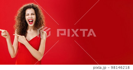 Woman feeling awesome on great party of friend pointing at entrance. Stylish carefree curly-haired female in red evening dress having fun near pool winking smiling joyfully and pointing left Woman feeling awesome on great party of friend pointing at entrance. Stylish carefree curly-haired female in red evening dress having fun near pool winking smiling joyfully and pointing left 98146218