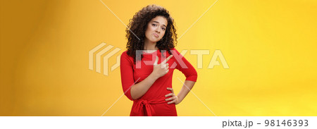 Upset disappointed and unimpressed attractive young european curly-haired woman in red dress pursing lips in gloomy face, frowning displeased as pointing at upper left corner from regret and dislike 98146393