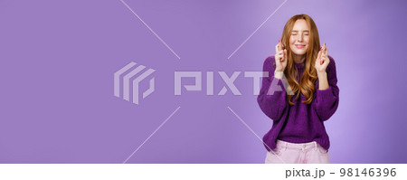 Girl makes wish eager dream come true. Atractive hopeful and positive young redhead woman in purple sweater close eyes and smiling as cross fingers for good luck, praying and anticipating miracle 98146396
