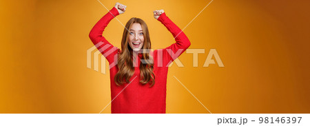 Optimistic happy and supportive ginger girl yelling cheering words, raising hands joyfully and smiling broadly triumphing, celebrating success and win, posing satisfied and excited against orange wall 98146397