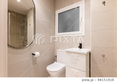Part of bathroom with toilet bowl, washbasin on table, beautiful oval mirror on wall lined with beige marble tiles, in which you can see reflection of shower area with metal fittings. Part of bathroom with toilet bowl, washbasin on table, beautiful oval mirror on wall lined with beige marble tiles, in which you can see reflection of shower area with metal fittings. 98151916