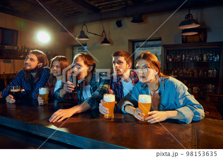 Group of young people, men and women, sport fans watching match at pub. Attentively looking, emotionally cheering up favourite team. 98153635