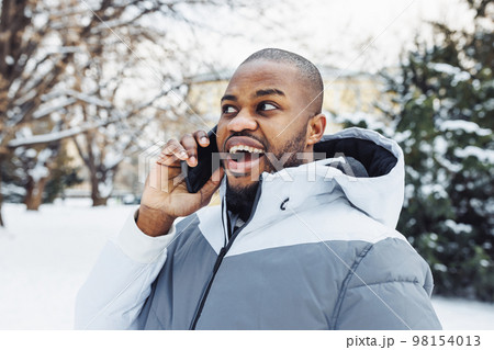 Happy surprised African American man in casual white sport clothes talking on phone, smiling and shocked in park or street outdoor. Winter season with snow 98154013