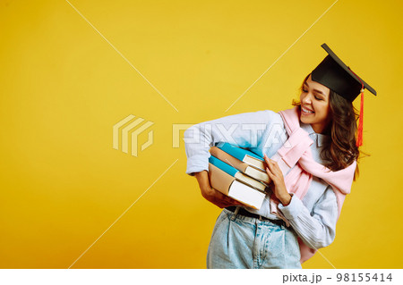 Graduate woman in a graduation hat on her head, with books stands on a yellow background. Graduate woman in a graduation hat on her head, with books stands on a yellow background. 98155414