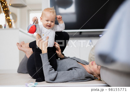 Happy family moments. Mother lying comfortably on children's mat playing with her baby boy watching and suppervising his first steps. Positive human emotions, feelings, joy Happy family moments. Mother lying comfortably on children's mat playing with her baby boy watching and suppervising his first steps. Positive human emotions, feelings, joy 98155771