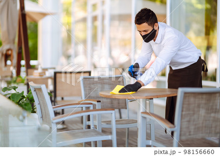Young Waiter in protective face mask and gloves sanitizing. cleaning the table with disinfectant 98156660