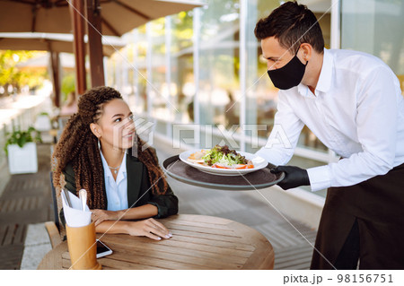 Young waiter in protective face mask and gloves while bringing food to a customer in cafe. Covid-19. 98156751