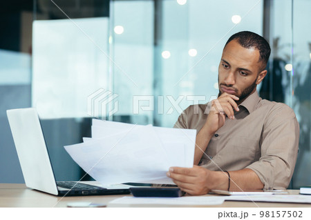 Pensive serious businessman reading financial report, hispanic businessman holding document in hands looking disappointed, working inside modern office with laptop behind paper work. 98157502