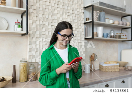 Happy and smiling young teenage girl standing at home in the kitchen and using the phone. Receives messages, chats with friends, boyfriend, chats news, blogs, social networks, online shopping. Happy and smiling young teenage girl standing at home in the kitchen and using the phone. Receives messages, chats with friends, boyfriend, chats news, blogs, social networks, online shopping. 98158361