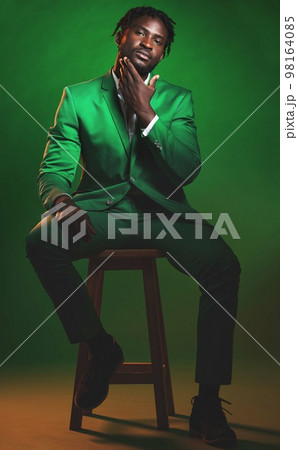Fashion, formal and portrait of a black man in a suit sitting on a chair in studio with a luxury outfit. Elegant, stylish and African male model with fashionable clothes isolated by green background. 98164085
