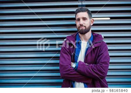 Handsome young man with hairstyle in fashionable clothes stands near iron wall and looking at the camera Handsome young man with hairstyle in fashionable clothes stands near iron wall and looking at the camera 98165840