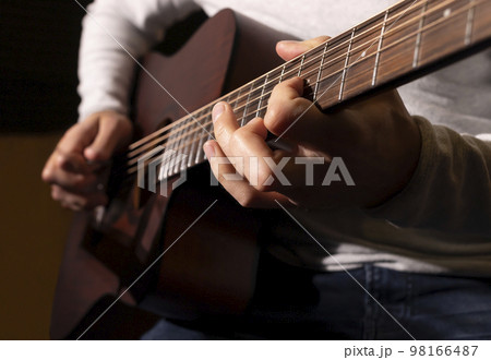 Closeup white man plays guitar, holding musical instrument in hands, sitting on chair in studio. Darken photo. Male wears casual cloth. Hobby, leisure of creative person, artist. Horizontal 98166487