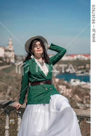 Woman walks around the city, lifestyle. A young beautiful woman in a green jacket, white skirt and hat is sitting on a white fence with balusters overlooking the sea bay and the city. Woman walks around the city, lifestyle. A young beautiful woman in a green jacket, white skirt and hat is sitting on a white fence with balusters overlooking the sea bay and the city. 98167920