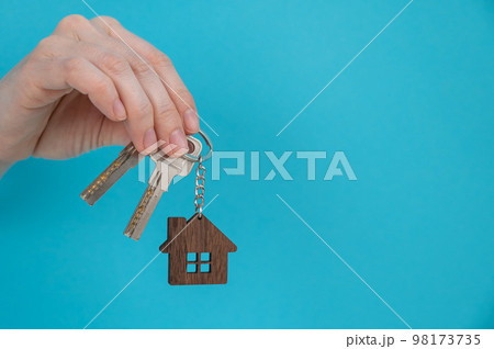 A woman's hand holds the keys with a keychain in the shape of a house on a blue background. A woman's hand holds the keys with a keychain in the shape of a house on a blue background. 98173735