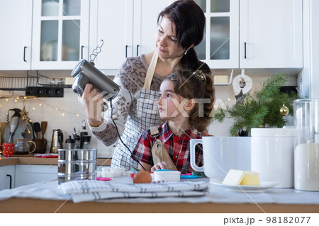 Mom and daughter in the white kitchen knead the dough in the bowl for gingerbread and cookies with a blender for Christmas and new year. 98182077