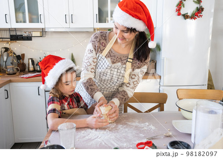 Mom and daughter in the white kitchen are preparing cookies for Christmas and new year. Family day, preparation for the holiday, learn to cook delicious pastries, cut shapes out of dough with molds Mom and daughter in the white kitchen are preparing cookies for Christmas and new year. Family day, preparation for the holiday, learn to cook delicious pastries, cut shapes out of dough with molds 98182097