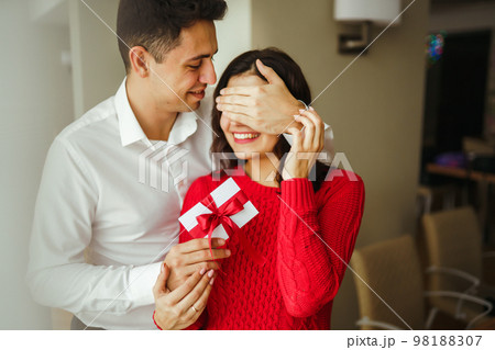 A young man gives his beloved a present and closes her eyes with a hand. Valentine's Day. 98188307