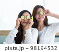 Home spa. Two beautiful young women place pieces of cucumber on their cheek in the home spa.. 98194532