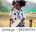 A cute little African-American girl in eyeglasses is sitting on a chair and holding a notepad and a pencil in a public Park on a beautiful morning.. 98194534