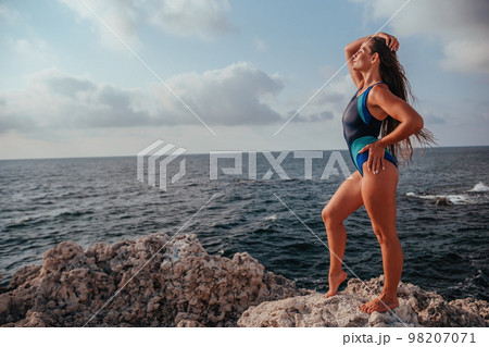 A young beautiful tanned woman with long hair in blue bikini stands at rock near blue green clear ocean sea water. Woman relax on sunset at sea, enjoy holidays and weekend vacation in summer time. A young beautiful tanned woman with long hair in blue bikini stands at rock near blue green clear ocean sea water. Woman relax on sunset at sea, enjoy holidays and weekend vacation in summer time. 98207071
