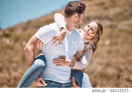 Young loving, affectionate and fun couple bonding while enjoying the day laughing outdoors in nature. Happy, in love and smiling man piggyback his wife while holding her outside on a valley or hill 98211073
