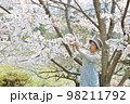 桜の枝を持ち上げて花を見ているシニア女性　ニーアップ 98211792