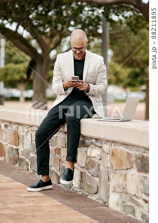 Stylish, cool smiling businessman on a phone in the park outside in nature. Happy man texting, chatting or reading social media messages on a smartphone outdoors on a break from work with copy space 98211895