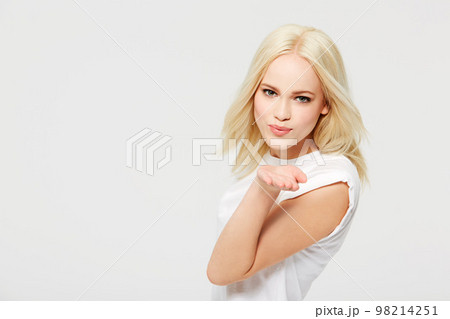 Young blond woman blowing a kiss with love and smile against a white studio background. Portrait of beautiful isolated casual model with blonde hair posing, flirting or kissing on mockup 98214251
