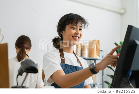 Asian woman cashier wears an apron and using pos terminal to input orders on coffee shop counter. 98215528