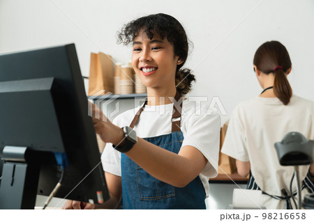 Asian woman cashier wears an apron and using pos terminal to input orders on coffee shop counter. 98216658