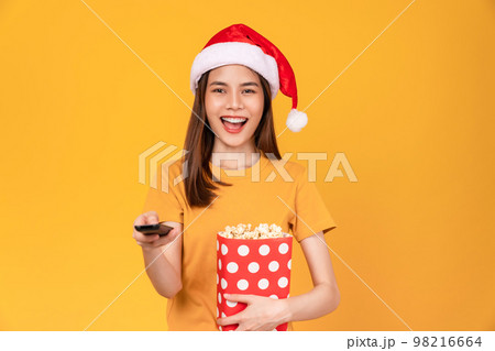 woman wearing Christmas hat with holding a red bucket of popcorn while the TV remote to watch movies 98216664