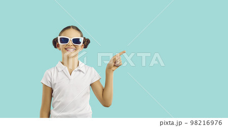 Caucasian girl teen in funny sunglasses with smile points finger at copy space in turquoise studio 98216976