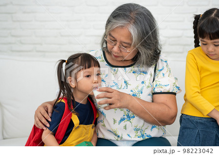 Happy elderly Asian Grandma sits beside her granddaughter and feeds fresh milk from glass for breakfast at home. Concept of a happy family and takes care together, preschool health care Happy elderly Asian Grandma sits beside her granddaughter and feeds fresh milk from glass for breakfast at home. Concept of a happy family and takes care together, preschool health care 98223042