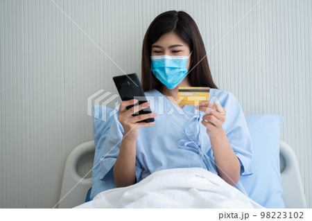 Happy Asian woman wearing a medical mask and holding mock up credit /insurance card and smartphone in a hospital bed for Check health insurance eligibility. Insurance policy by bank, payment medical Happy Asian woman wearing a medical mask and holding mock up credit /insurance card and smartphone in a hospital bed for Check health insurance eligibility. Insurance policy by bank, payment medical 98223102