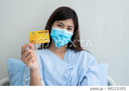 Happy Asian woman wearing a medical mask and holding mock up credit card /insurance card and ok sign in a hospital bed. Insurance policy by bank, payment medical treatment concept 98223103