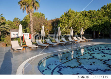 Empty deck chairs and folded parasols surrounded with trees arranged at poolside with clear blue sky in the background at tourist resort Empty deck chairs and folded parasols surrounded with trees arranged at poolside with clear blue sky in the background at tourist resort 98226139