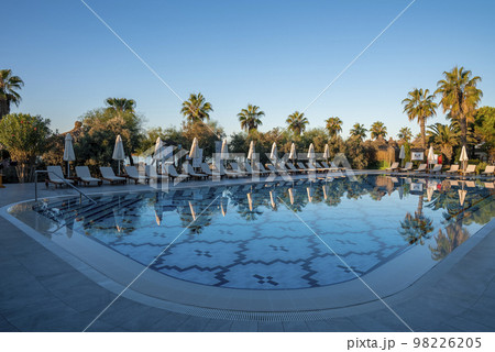Empty deck chairs and folded parasols surrounded with trees arranged in a row at poolside with clear blue sky in the background at tourist resort 98226205