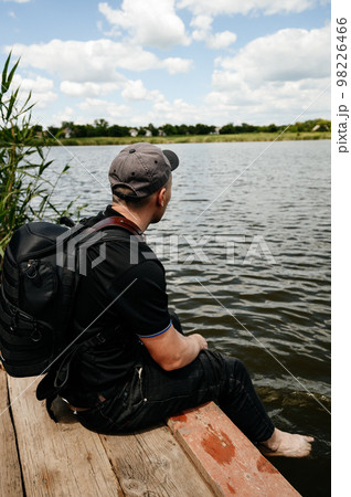 A man sits on the edge of a wooden bridge near a lake next to reeds. A man sits on the edge of a wooden bridge near a lake next to reeds. 98226466