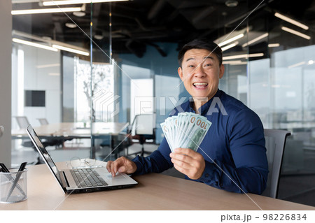 A young Asian businessman is sitting in the office at a table with a laptop. Holds cash money in hands. Plays online games, places bets, checks account, successful deal, investment. He looks at the 98226834