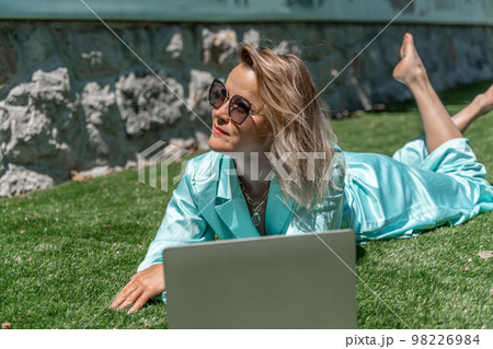 a young beautiful woman with blond curly hair in glasses and a blue dress sits on the grass in nature and uses a laptop, 98226984