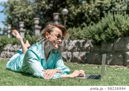 a young beautiful woman with blond curly hair in glasses and a blue dress sits on the grass in nature and uses a laptop, 98226994