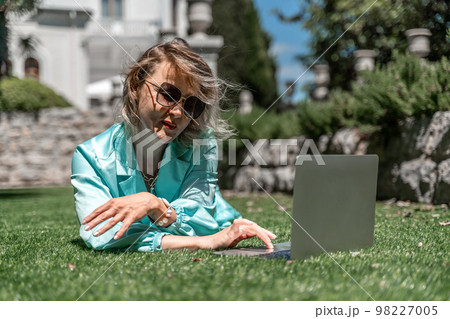 a young beautiful woman with blond curly hair in glasses and a blue dress sits on the grass in nature and uses a laptop, 98227005