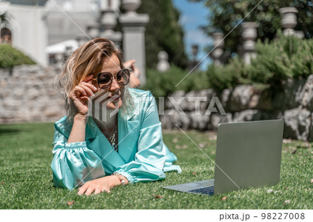 a young beautiful woman with blond curly hair in glasses and a blue dress sits on the grass in nature and uses a laptop, 98227008