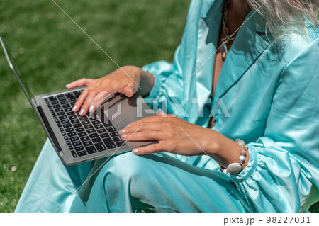 a young beautiful woman with blond curly hair in glasses and a blue dress sits on the grass in nature and uses a laptop, 98227031