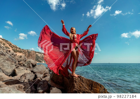 Red dress sea woman. A blonde with flowing hair in a long flowing red dress stands on a rock near the sea. Travel concept, photo session at sea Red dress sea woman. A blonde with flowing hair in a long flowing red dress stands on a rock near the sea. Travel concept, photo session at sea 98227184