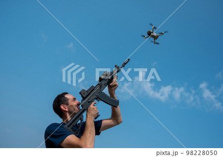 A man aims to shoot a rifle at a flying drone against a blue sky.  98228050